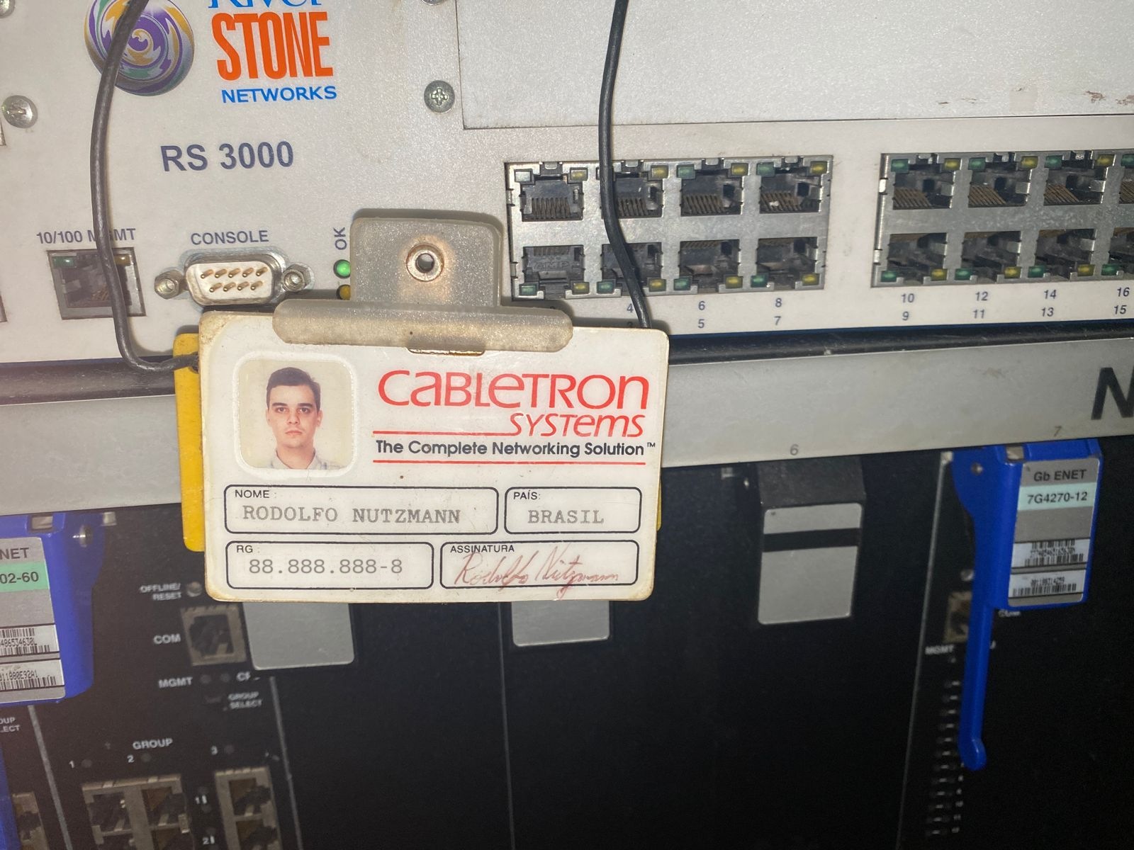 A 1996 Cabletron Systems photo identity badge for Rodolfo Nutzmann, Brazil, hanging by its clip from the front panel of a Riverstone Networks RS 3000 router. The identity-document number has been replaced with repeated digits.