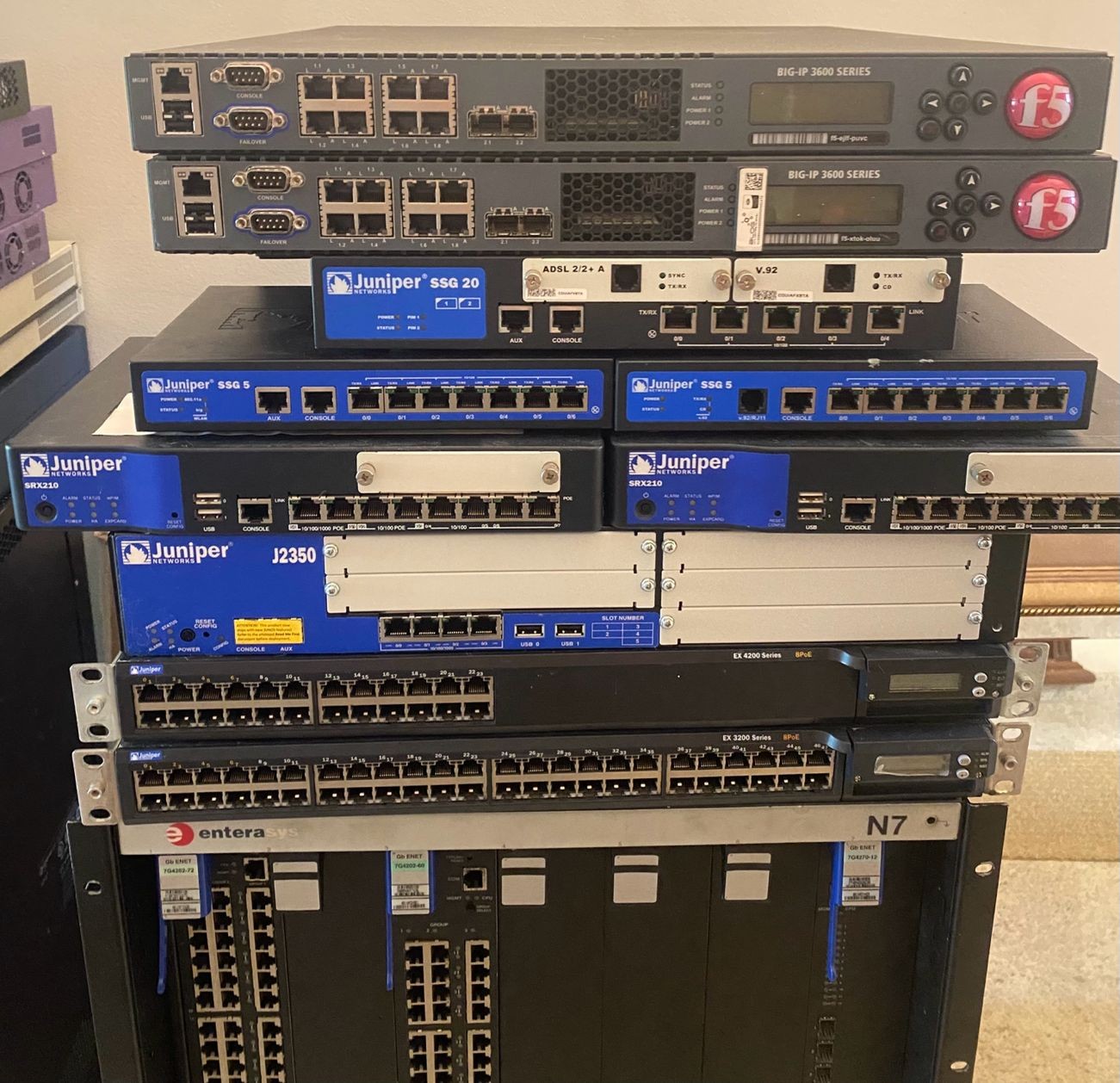 A home lab stack of decommissioned network appliances: two F5 BIG-IP 3600 units, a Juniper SSG 20, two Juniper SSG 5 firewalls, two SRX210 gateways, a J2350 router, EX4200 and EX3200 switches, and an Enterasys N7 chassis underneath.